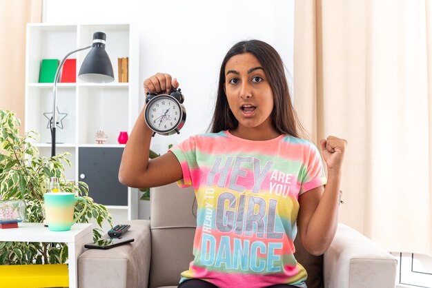 Young girl in casual clothes holding alarm clock clenching fist happy and excited sitting on the chair in light living room