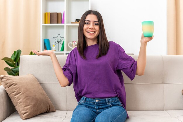 Young girl in casual clothes holding cup of tea smiling cheerfully looking presenting something with arm of hand sitting on a couch in light living room