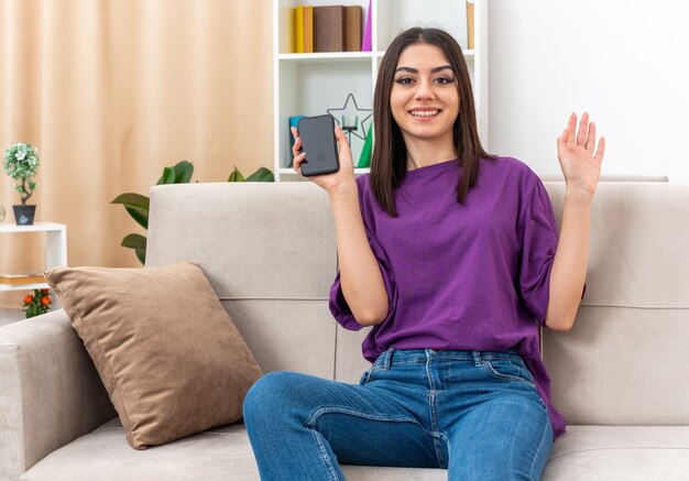 Young girl in casual clothes holding smartphone looking at camera happy and positive smiling waving with hand sitting on a couch in light living room