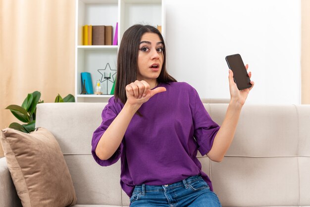 Young girl in casual clothes holding smartphone looking confused pointing with thumb at mobile sitting on a couch in light living room