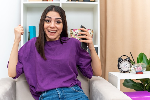 Young girl in casual clothes holding smartphone looking happy and excited clenching fist sitting on a chair in light living room