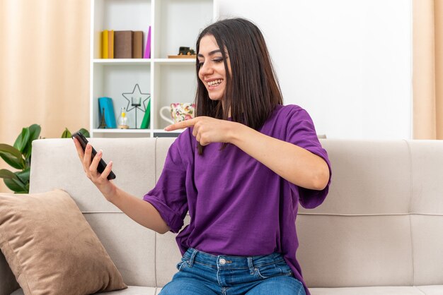 Young girl in casual clothes holding smartphone pointing with index finger at it happy and cheerful smiling broadly sitting on a couch in light living room