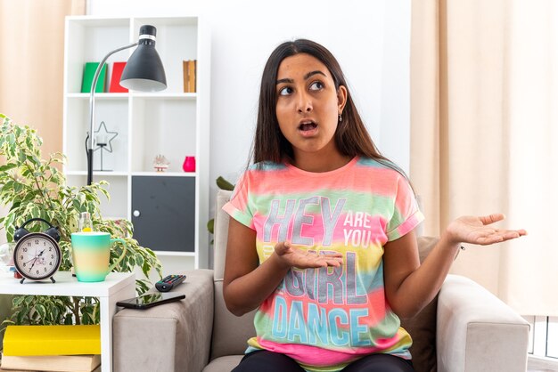 Young girl in casual clothes looking aside confused presenting with arm of hands sitting on the chair in light living room