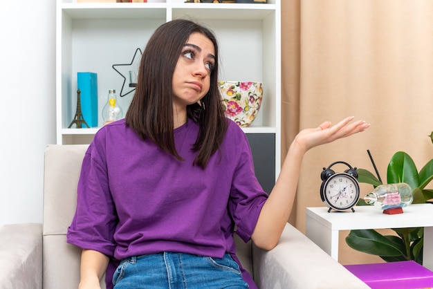 Young girl in casual clothes looking aside with sad expression presenting something with arm of her hand sitting on a couch in light living room