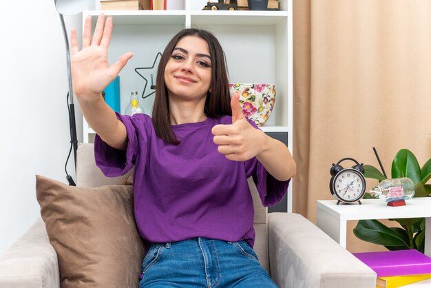 Young girl in casual clothes looking happy and positive showing number five and thumbs up sitting on a chair in light living room