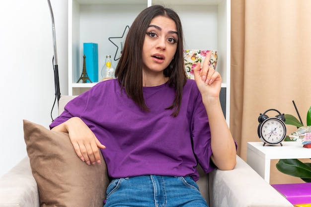 Young girl in casual clothes looking happy and surprised showing index finger having new idea sitting on a chair in light living room