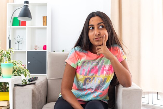 Young girl in casual clothes looking up puzzled sitting on the chair in light living room