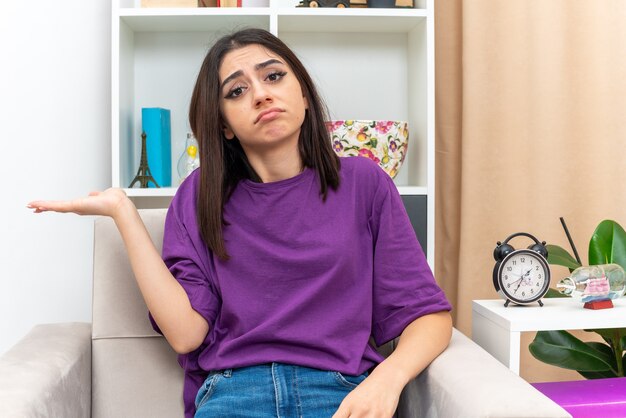 Young girl in casual clothes looking  with sad expression on face presenting something with arm of her hand sitting on a chair in light living room