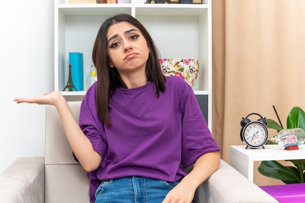 Young girl in casual clothes looking  with sad expression on face presenting something with arm of her hand sitting on a chair in light living room