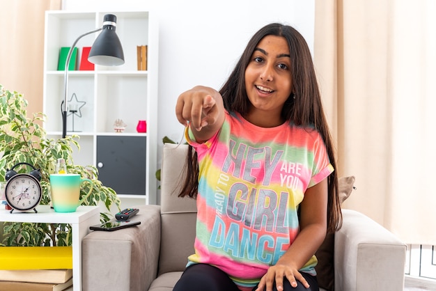 Young girl in casual clothes pointing with index finger  smiling cheerfully happy and positive sitting on the chair in light living room