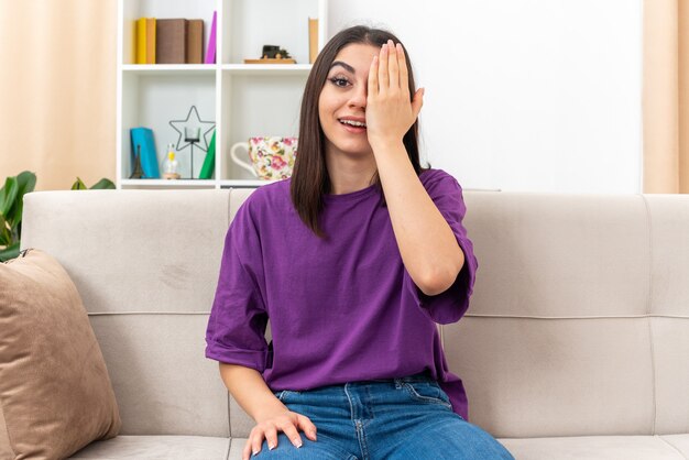 Young girl in casual clothes smiling cheerfully covering one eye with hand sitting on a couch in light living room