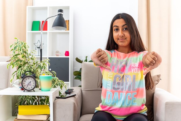 Young girl in casual clothes  smiling confident showing thumbs down sitting on the chair in light living room