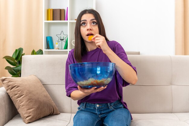 Young girl in casual clothes with bowl of chips eating and looking intrigued sitting on a couch in light living room