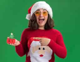 Free photo young girl in christmas sweater wearing santa hat and glasses holding toy cubes with new year date pointing with index finger at it smiling cheerfully happy and excited standing over green wall