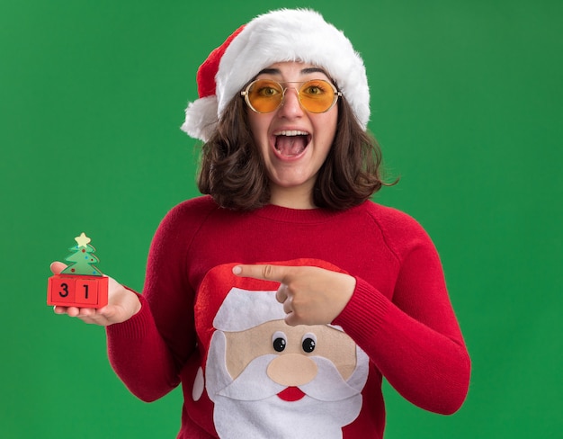 Free Photo young girl in christmas sweater wearing santa hat and glasses holding toy cubes with new year date pointing with index finger at it smiling cheerfully happy and excited standing over green wall