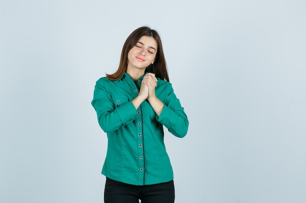 Young girl clasping hands over chest in green blouse, black pants and looking cheery. front view.