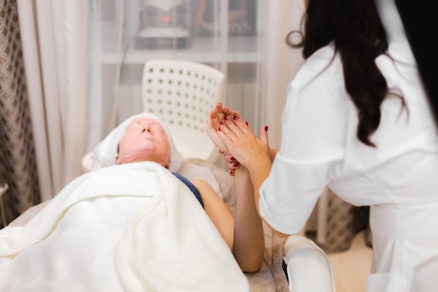 A young girl client with an alginate mask on her face lies on the cosmetology table, the beautician makes a manual hand massage