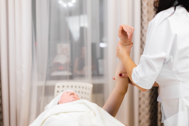 A young girl client with an alginate mask on her face lies on the cosmetology table, the beautician makes a manual hand massage