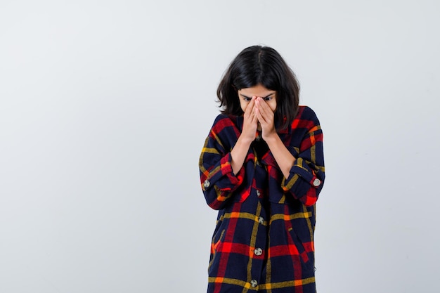 Young girl covering mouth and nose with hands in checked shirt and looking surprised. front view.