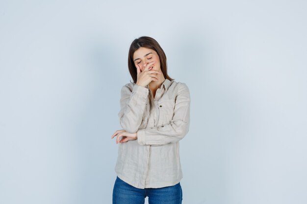 Young girl covering mouth while laughing in beige shirt, jeans and looking cheery , front view.