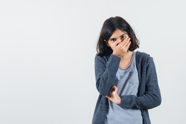 Young girl covering mouth with hand in light gray t-shirt and dark grey zip-front hoodie and looking surprised.