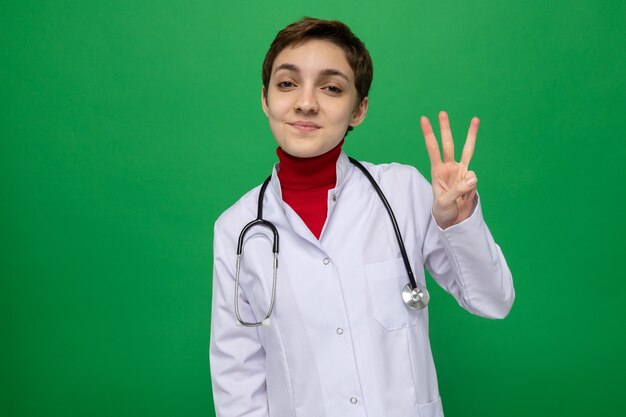 Young girl doctor in white coat with stethoscope around neck looking at front happy and positive smiling confident showing number three with fingers standing over green wall