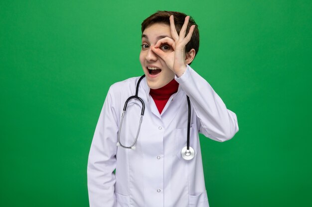 Young girl doctor in white coat with stethoscope around neck looking through fingers making ok sign smiling cheerfully standing on green