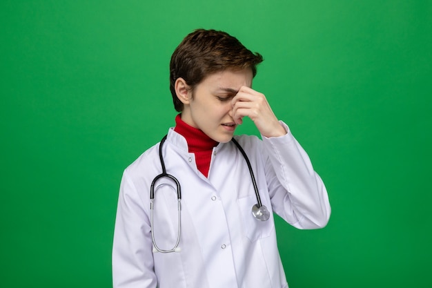 Young girl doctor in white coat with stethoscope around neck looking unwell tired and onworked touching ner nose between closed eyes standing on green