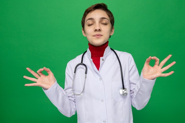 Young girl doctor in white coat with stethoscope making meditation gesture trying to relax with eyes closed