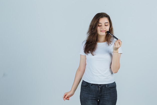 Young girl doing makeup with brush in t-shirt, jeans and looking charming , front view.