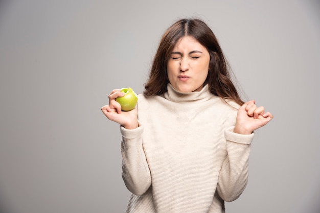 Free photo a young girl eating a green apple on a gray wall.