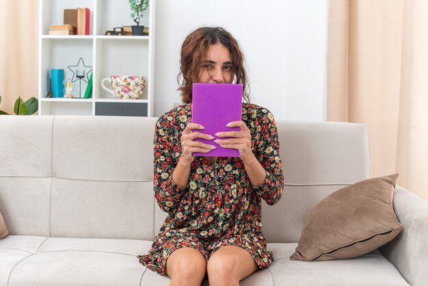 Young girl in floral dress holding book looking happy and positive sitting on a couch in light living room