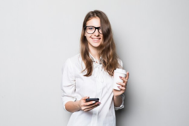 Young girl in glasses dressed up in strict office white t-shirt stands in front of white wall and holds her phone and coffee cup in hands