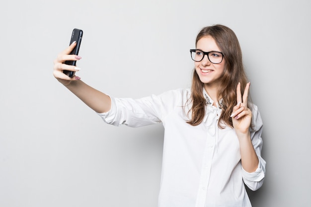 Young girl in glasses dressed up in strict office white t-shirt stands in front of white wall and holds her phone in hands