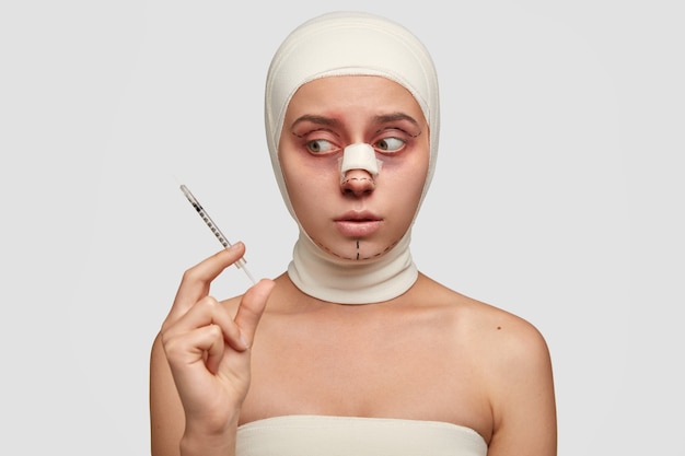 Free photo young girl going to have skin injection, looks at needle with anodyne serum in embarrassement, has marked dotted lines on face, has bandage on bridge of nose after otoplasty procedure