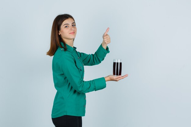 Young girl in green blouse, black pants holding glass of black liquid, showing thumb up and looking cheery , front view.