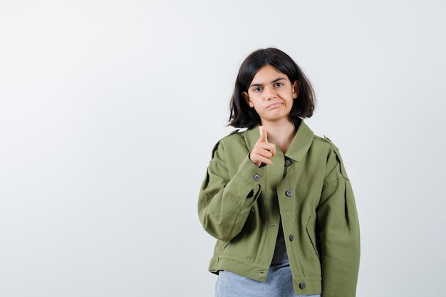 Young girl in grey sweater, khaki jacket, jean pant pointing and looking serious , front view.