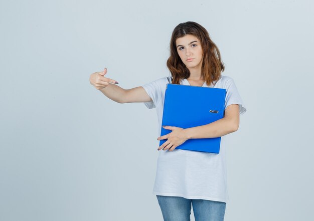 Young girl holding folder, pointing to the right side in white t-shirt and looking hesitant. front view.