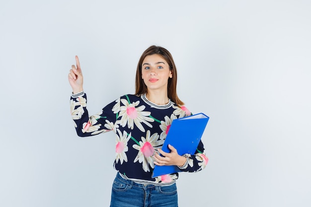 Young girl holding folder, pointing up in floral blouse, jeans and looking curious , front view.