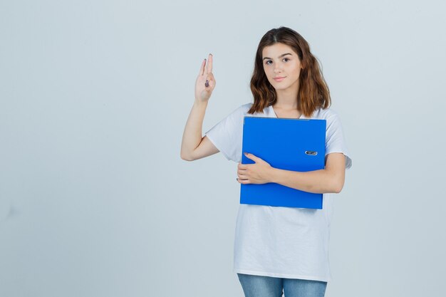 Young girl holding folder, showing ok gesture in white t-shirt and looking jolly , front view.