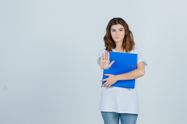 Young girl holding folder, showing stop gesture in white t-shirt and looking confident , front view.