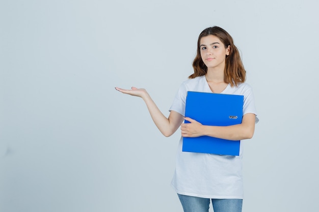 Young girl holding folder, showing welcoming gesture in white t-shirt and looking confident , front view.