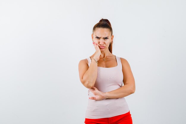 Young girl holding hand near mouth, grimacing while posing in beige top and red pants and looking serious , front view.