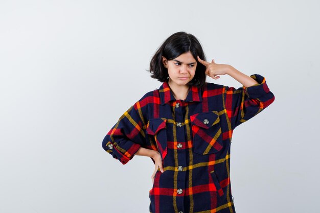 Young girl holding hand on waist, putting index finger on temple in checked shirt and looking pensive , front view.