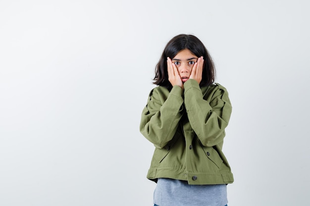 Young girl holding hands on cheeks in grey sweater, khaki jacket, jean pant and looking surprised. front view.