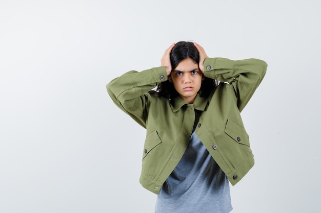 Free photo young girl holding hands on head in grey sweater, khaki jacket, jean pant and looking serious , front view.