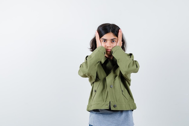 Young girl holding hands on head in grey sweater, khaki jacket, jean pant and looking serious , front view.