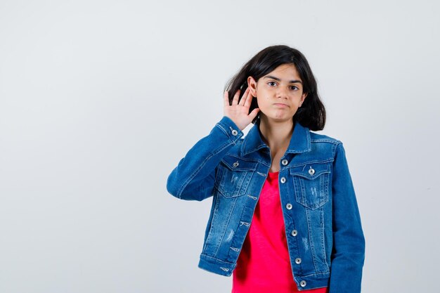 Young girl holding hands near ear to hear something in red t-shirt and jean jacket and looking focused, front view.