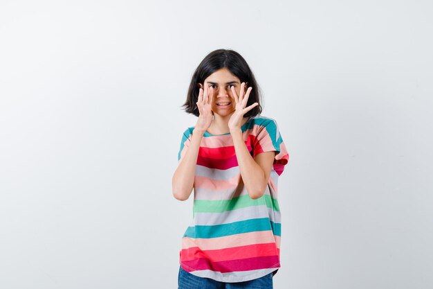 Young girl holding hands near mouth as calling someone in colorful striped t-shirt and looking happy , front view.