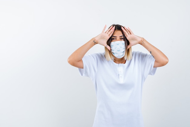 Young girl holding hands on temples to see clearly in white t-shirt and mask and looking focused , front view.